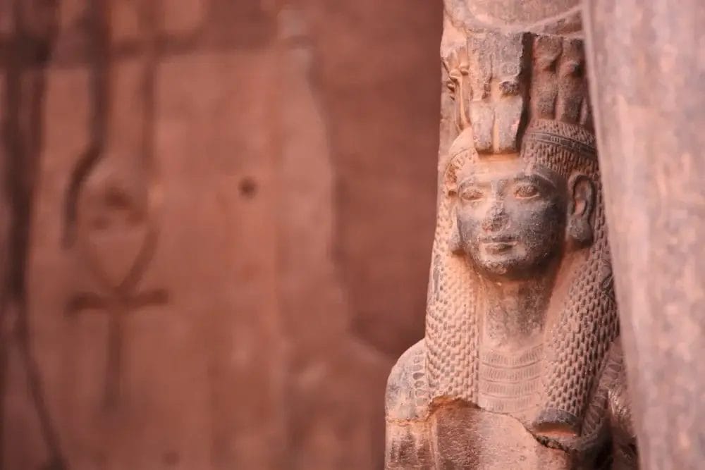 Primo piano di un'antica statua egizia in pietra di una donna con un copricapo dettagliato e capelli intrecciati, simile a una bozza automatica, con un'incisione murale sbiadita di un simbolo ankh visibile sullo sfondo sfocato.