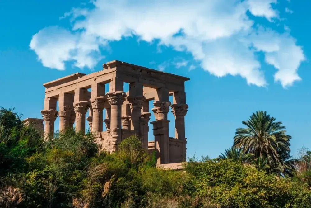 Un antico tempio in pietra con alte colonne si erge su una collina verdeggiante, circondato da palme sotto un cielo azzurro con nuvole bianche, immortalato in una splendida bozza automatica.