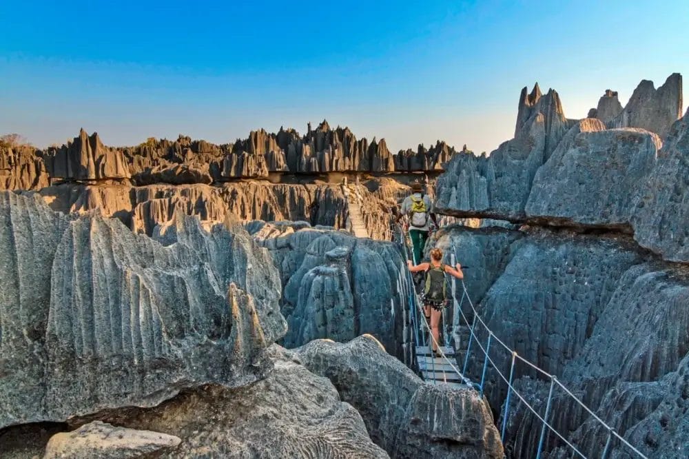 Due persone attraversano uno stretto ponte sospeso su pinnacoli di calcare grigio frastagliato in un paesaggio roccioso e drammatico, sotto un cielo azzurro e limpido, nel Parco nazionale Tsingy de Bemaraha, in Madagascar.