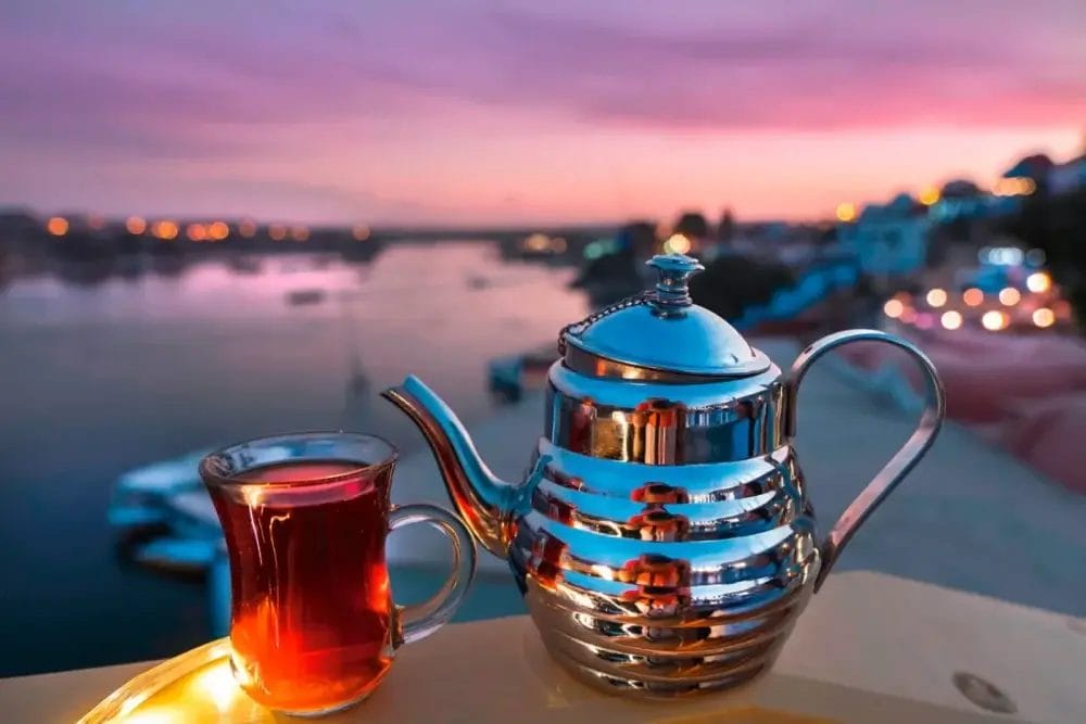Un bicchiere di tè e una teiera di metallo lucido si trovano su un cornicione che si affaccia su un fiume al tramonto, con le luci sfocate della città e i colori pastello del cielo che si riflettono sull'acqua, creando una tranquilla atmosfera da Bozza automatica.