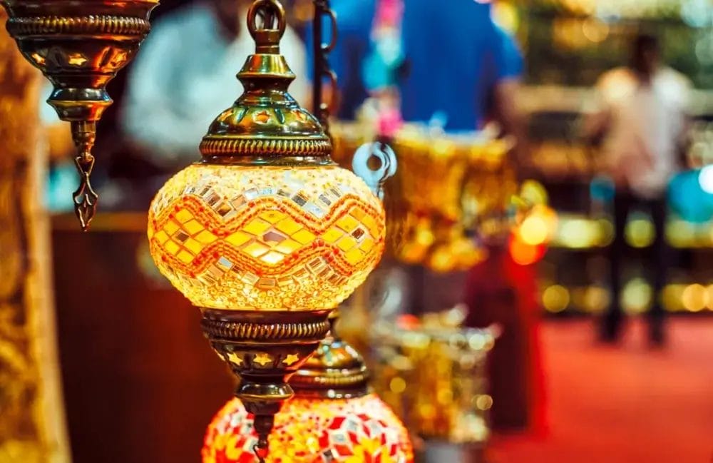 Lanterne a mosaico colorate e decorate con intricate lavorazioni metalliche sono appese in fila in un mercato. Lo sfondo, leggermente sfocato, mette in risalto il caldo bagliore e i motivi vivaci di queste lampade, creando una scena che ricorda una Bozza automatica.