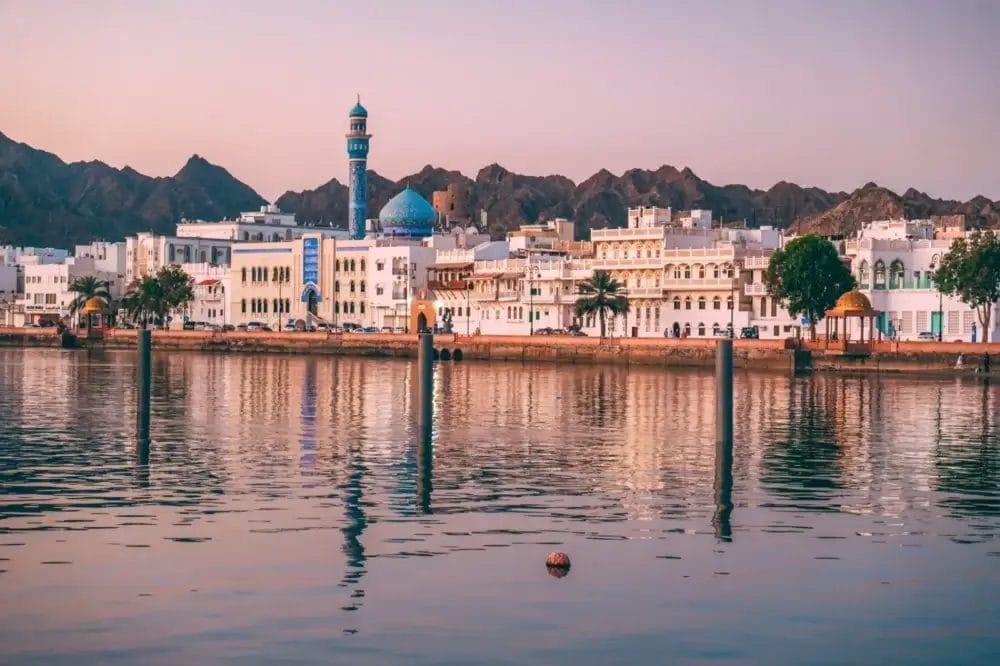 Veduta del lungomare di Muscat, Oman, con edifici bianchi e una moschea dalle cupole blu, sullo sfondo di montagne rocciose al tramonto; l'acqua calma riflette il paesaggio urbano e il cielo pastello, creando un'atmosfera serena e automatica di Bozza.