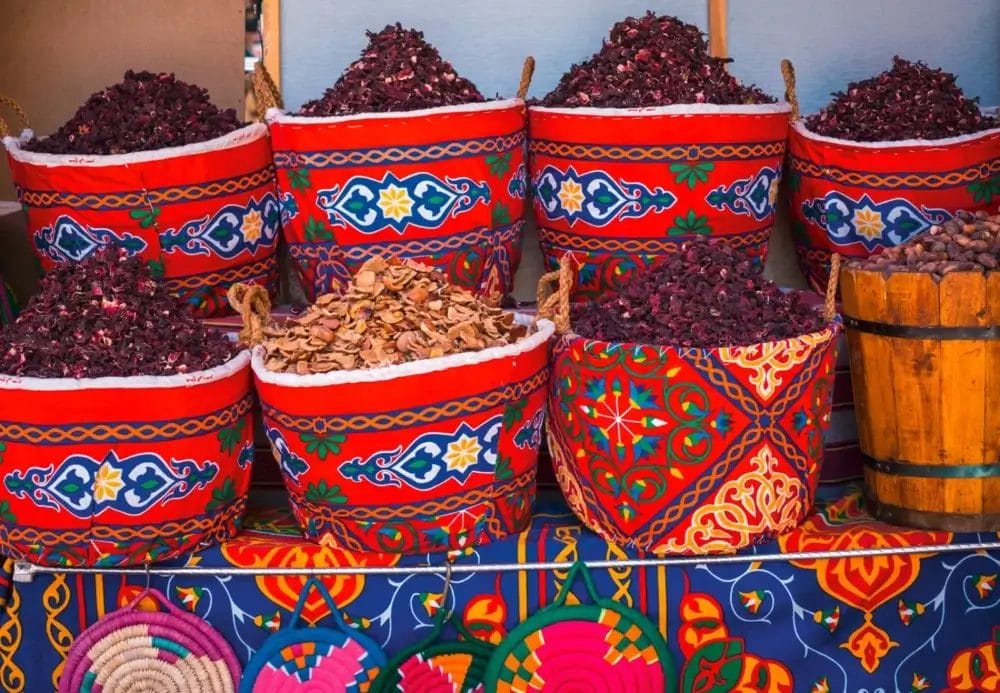 Su una vivace bancarella, che ricorda i mercati lungo il Nilo, sono esposti cestini colorati e fantasia, pieni di erbe o petali essiccati. Un contenitore di legno con piccoli pezzi marroni si trova sul lato destro, evocando le antiche tradizioni Piramidi.