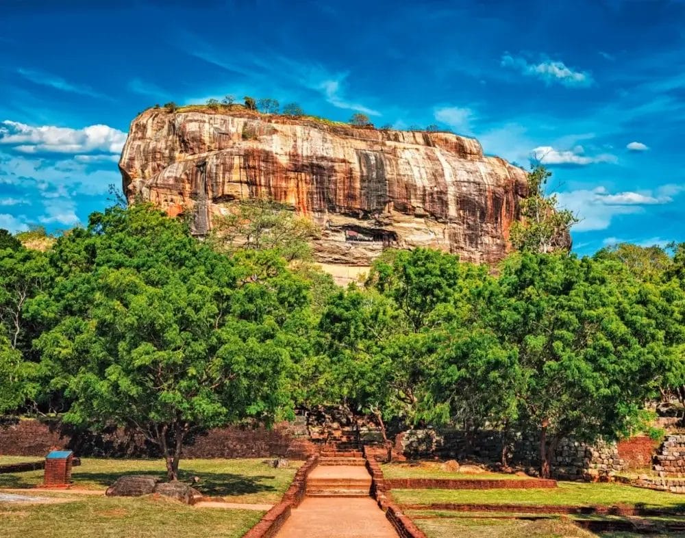 Una grande formazione rocciosa dalla cima piatta si erge al di sopra di alberi verdi e di un sentiero di pietra, sotto un cielo blu brillante con nuvole sparse. La scena, rigogliosa e vibrante, emana un forte spirito di natura, con erba verde e rovine di mattoni in primo piano.