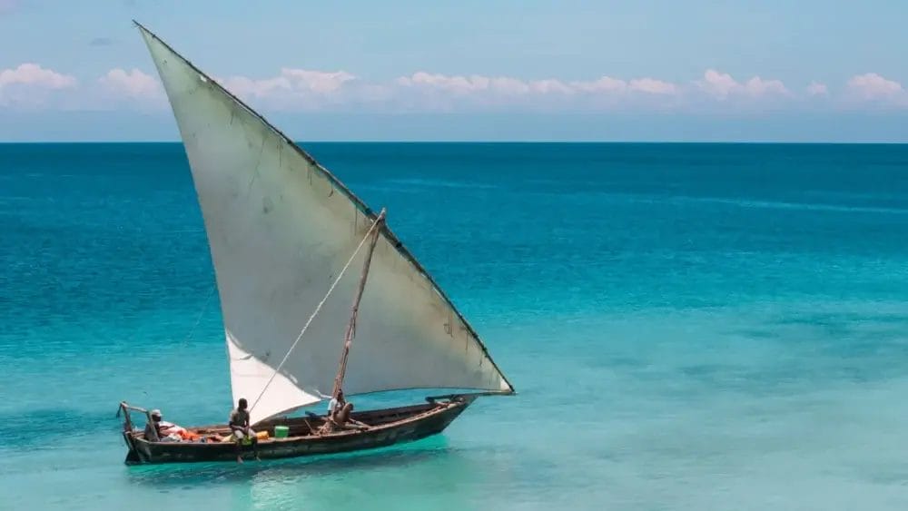 Una piccola barca a vela in legno con una grande vela bianca triangolare scivola sull'acqua limpida e turchese sotto un cielo azzurro. Diverse persone sono sulla barca e si godono il momento di pace, come una bozza automatica attraverso l'orizzonte calmo.