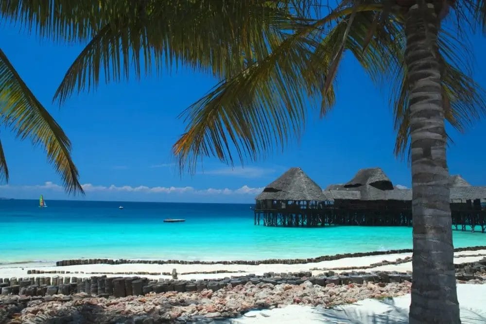 Una scena di spiaggia tropicale con sabbia bianca, acqua limpida e turchese e una capanna con tetto di paglia su palafitte a picco sul mare. Le foglie delle palme incorniciano l'immagine sotto un cielo blu brillante, creando un'ambientazione perfetta per una fuga automatica di Bozza.