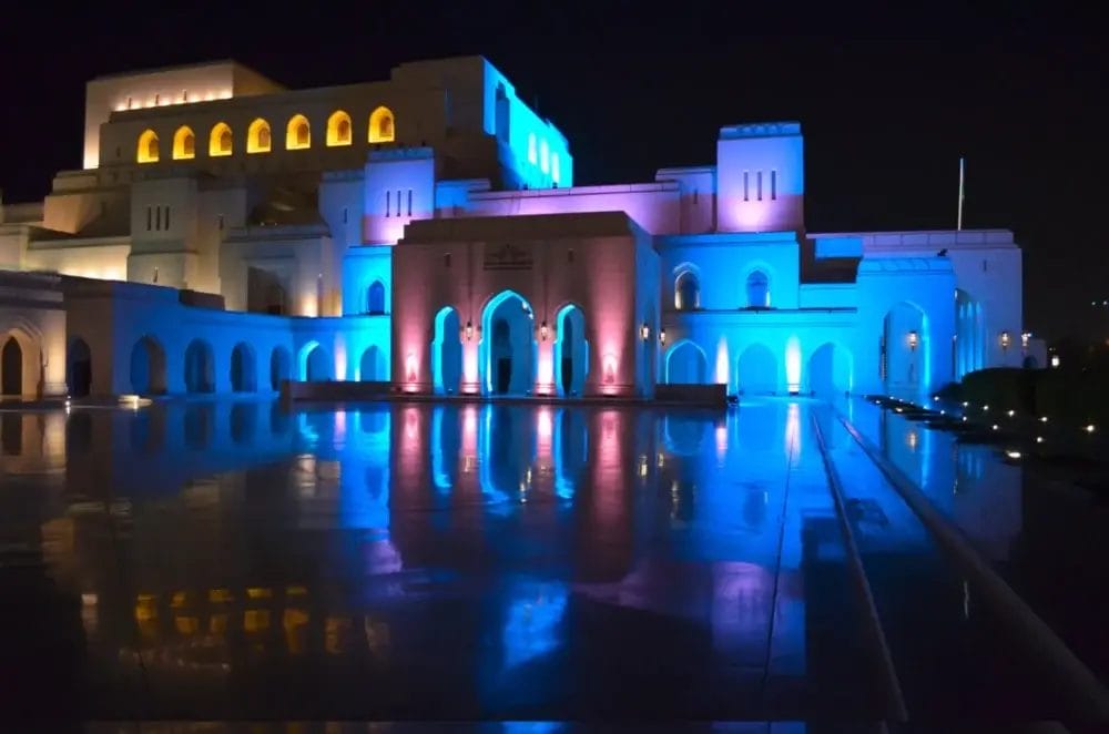 Un grande edificio bianco illuminato di notte con luci blu e rosa che si riflettono su una superficie lucida. La struttura presenta archi, colonne e più livelli con finestre luminose: una scena impressionante che ricorda una bozza automatica.