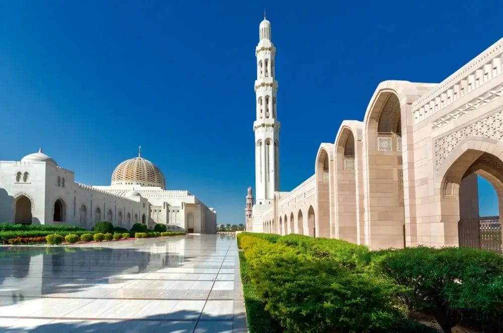 L'immagine mostra la Grande Moschea del Sultano Qaboos a Muscat, in Oman, con l'alto minareto, la cupola ornata, i camminamenti in marmo bianco e i corridoi ad arco sotto un cielo azzurro e limpido: un vero capolavoro di Bozza automatica incorniciato da cespugli verdeggianti.