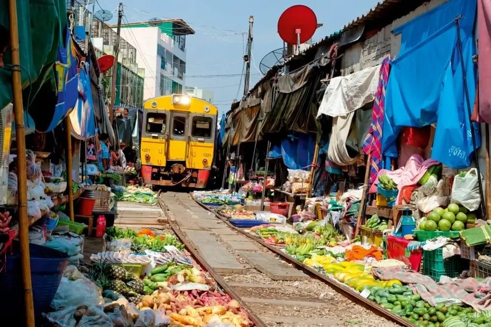 Un treno giallo si muove lentamente attraverso una stretta strada del mercato di Thailandia, fiancheggiata da frutta, verdura e ombrelli colorati: una scena indimenticabile per ogni amante del turismo che esplora la magia di Thailandia Magica.