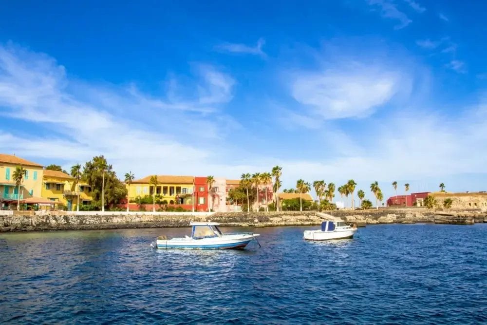 Due piccole imbarcazioni galleggiano sull'acqua azzurra vicino a un litorale con edifici colorati, palme e un muro di pietra, il tutto sotto un cielo azzurro brillante con nuvole a batuffolo: una scena che ricorda una perfetta bozza automatica.
