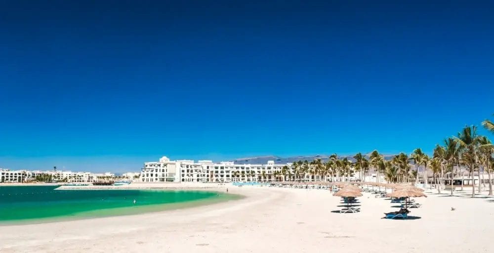 Una spiaggia incontaminata di sabbia bianca con acqua limpida e turchese, file di palme, ombrelloni di paglia, sedie a sdraio e un grande edificio bianco del resort creano una Bozza automatica di relax sotto un cielo blu brillante.