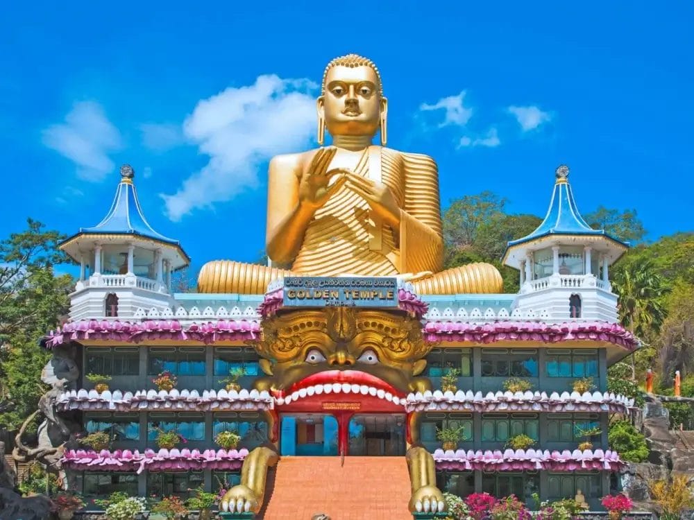 Una grande statua di Buddha dorata si trova in cima al colorato Tempio d'Oro di Dambulla, che cattura l'essenza dello Sri Lanka tra decorazioni ornamentali, un cielo blu e una vegetazione lussureggiante sullo sfondo.