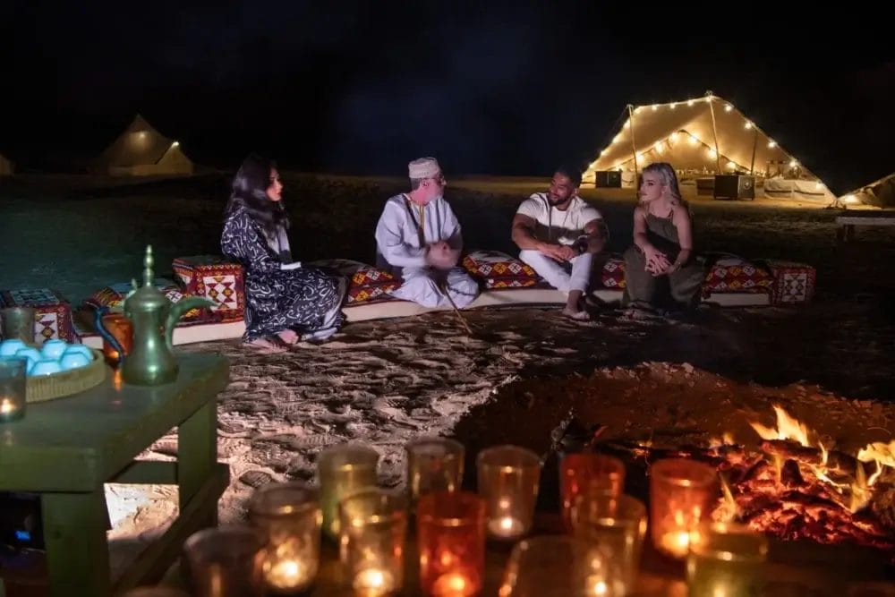 Quattro persone sedute su cuscini attorno a un fuoco di notte, parlano e si rilassano. Una tenda decorata con luci a corda fa da sfondo, mentre candele, una teiera e un tocco di Bozza automatica creano un'accogliente ambientazione da campo nel deserto.