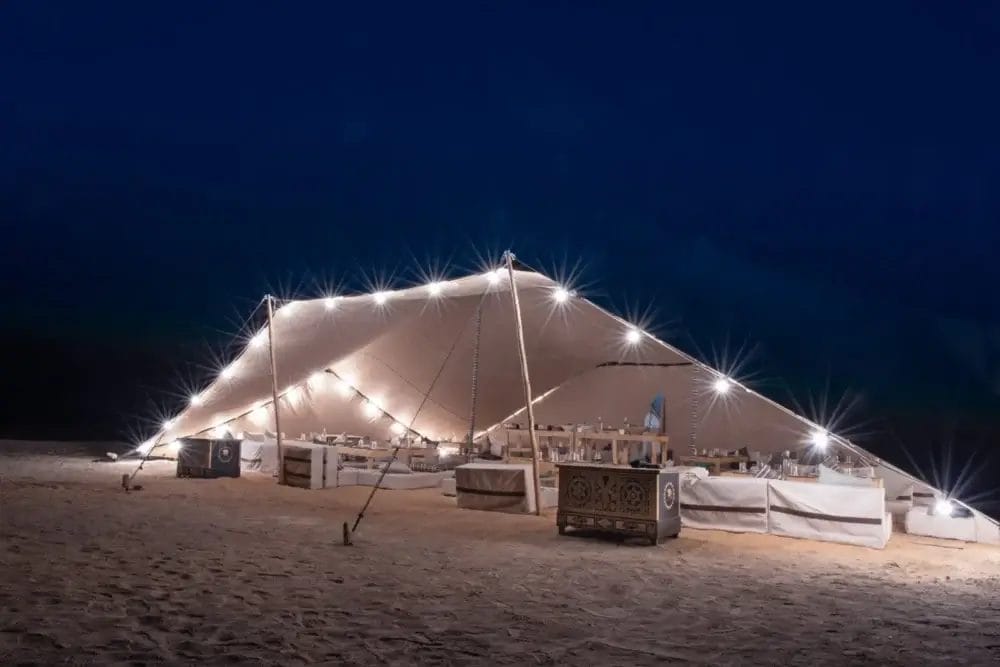 Una grande tenda illuminata con luci a corda si trova su un terreno sabbioso di notte, al riparo di un lungo tavolo da pranzo apparecchiato per un evento formale. Il cielo è scuro e la scena di festa ha il fascino di una bozza automatica, calda e invitante.