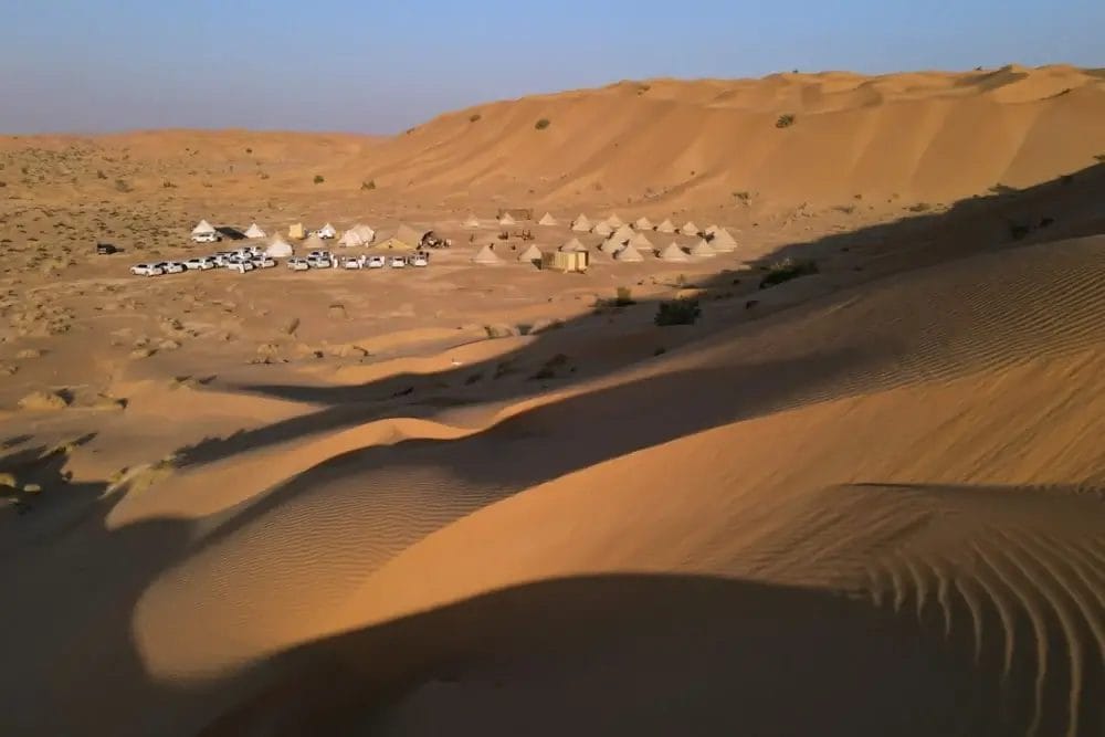 Un paesaggio desertico con file di tende bianche e diversi veicoli disposti alla base di alte dune di sabbia dorata sotto un cielo azzurro e limpido, ripreso da Bozza automatica. Le ombre delle dune si allungano sulla sabbia.