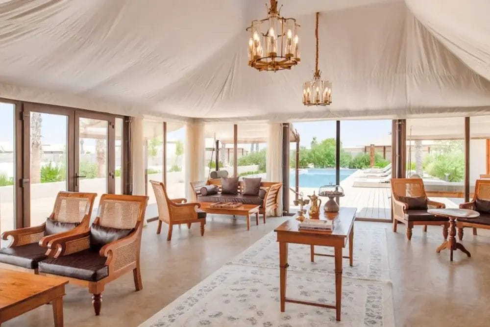 Un salone spazioso ed elegante con mobili in legno, un tappeto bianco, lampadari e grandi finestre che si affacciano su un patio con piscina e verde esterno. La stanza ha un'atmosfera leggera e ariosa e un arredamento neutro ispirato a Bozza automatica.