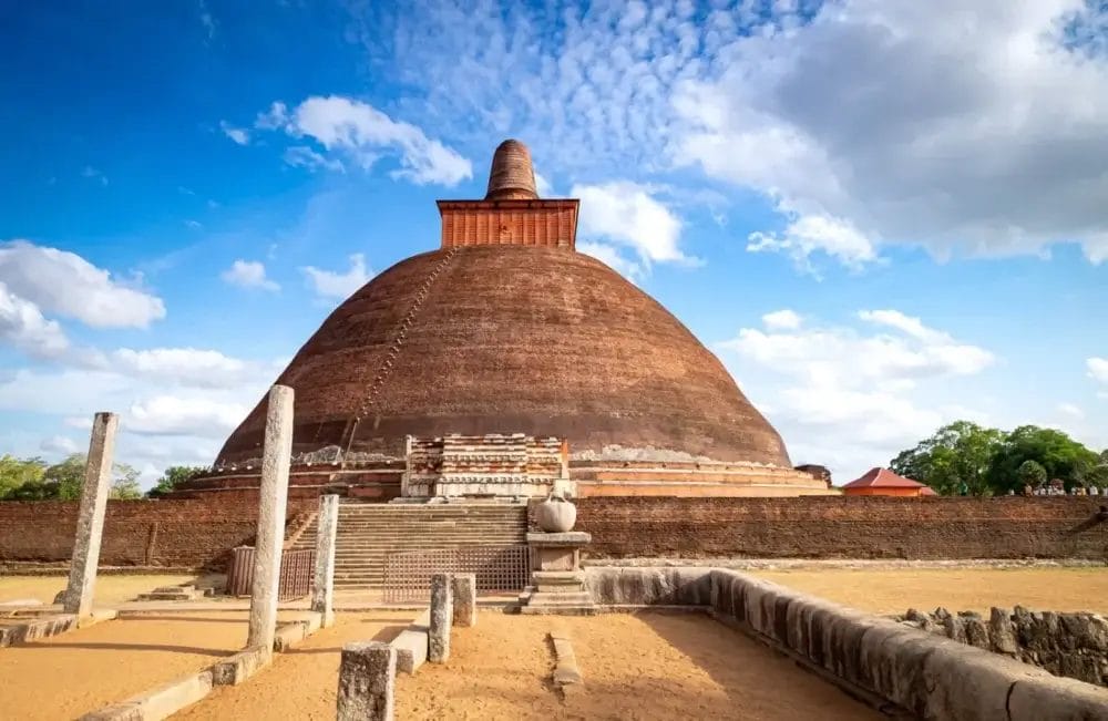 Un grande stupa antico fatto di mattoni rossi, circondato da colonne di pietra e rovine sotto un cielo blu con nuvole sparse. Questo sito storico su un terreno sabbioso, con gradini che salgono, cattura l'Essenza senza tempo dello Sri Lanka.