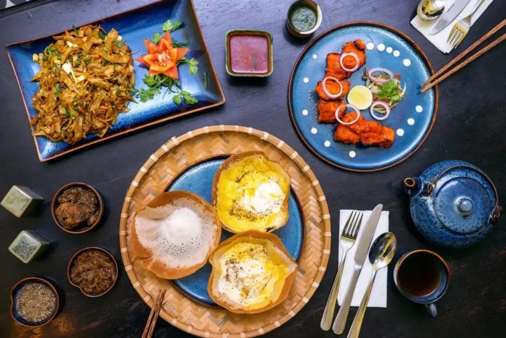 Una tavola imbandita per il viaggio in Sri Lanka, con piatti dello Sri Lanka come uova, noodles saltati in padella, pollo fritto, chutney, salse, una teiera e una tazza, splendidamente disposti su una superficie di legno scuro con bacchette e posate.