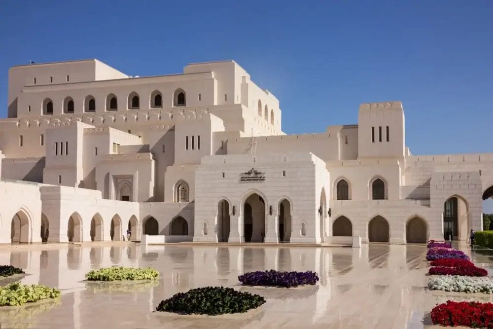L'immagine mostra la Royal Opera House Muscat, un grande edificio in pietra bianca con finestre e ingressi ad arco. Aiuole colorate decorano il cortile lucido, catturando l'eleganza di Muscat e Mare sotto un cielo azzurro e limpido.