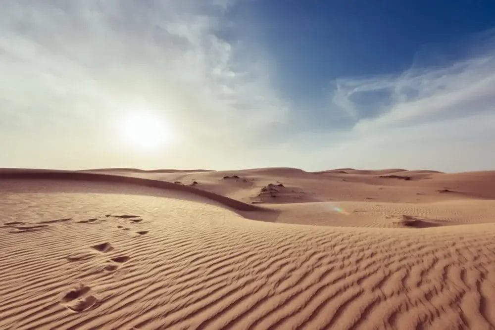 Dune di sabbia increspate si estendono in lontananza sotto un sole splendente e un cielo azzurro con nuvole sottili. In mezzo al paesaggio desertico, alcune impronte sparse e piccole rocce accennano a un viaggio, come una bozza automatica creata dalla natura.