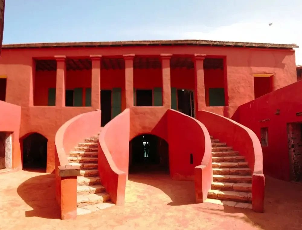 Un edificio simmetrico di colore rosso con due scale curve in pietra che conducono a un balcone, sullo sfondo di un cielo azzurro. La struttura presenta porte e finestre verdi, un ingresso ad arco sotto le scale ed emana il fascino di Bozza automatica.