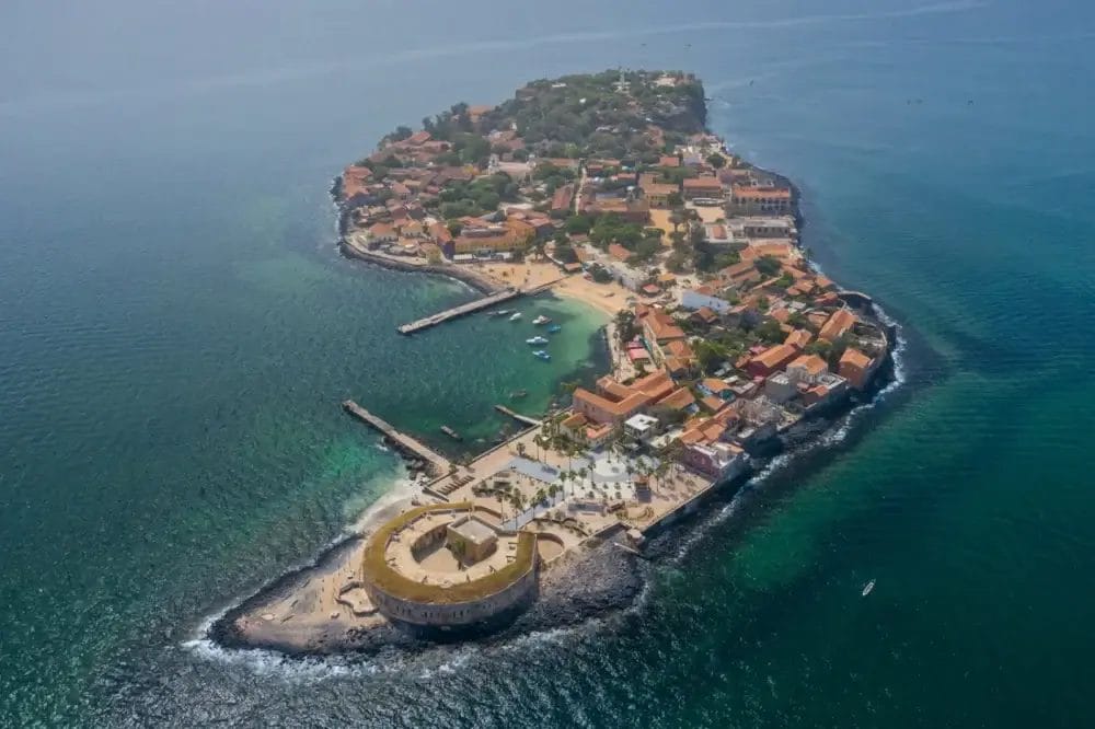 Veduta aerea dell'isola di Gorée, in Senegal, circondata da un oceano verde-blu, con edifici storici, moli e il Fort d'Estrées in pietra rotonda sulla punta dell'isola, una scena che ricorda una Bozza automatica.