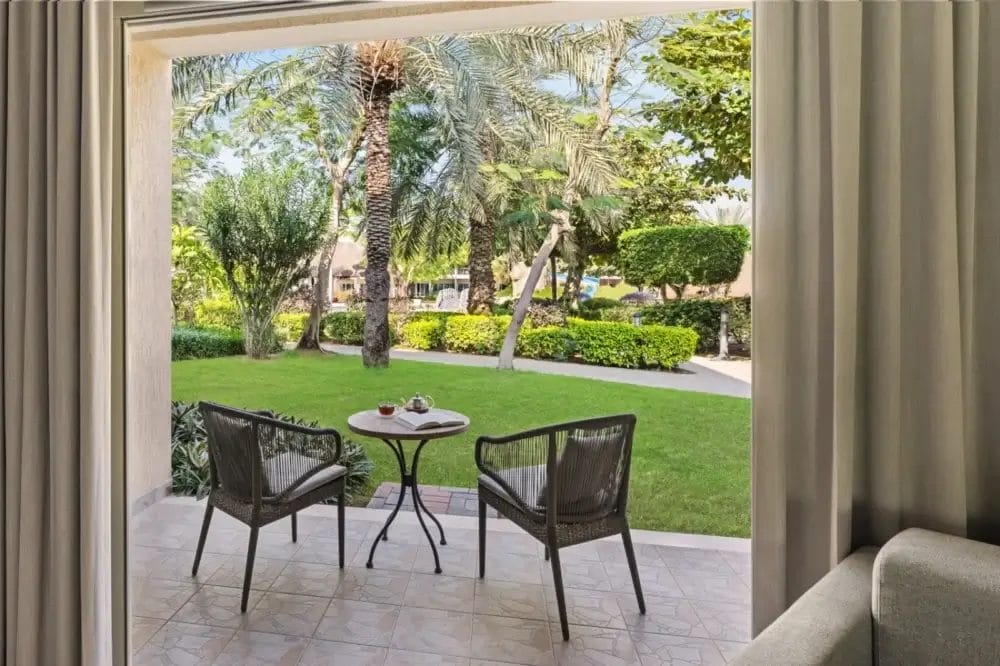 Un accogliente patio con due sedie di vimini e un tavolino rotondo si affaccia su un giardino verde e rigoglioso, con palme e cespugli curati: un rifugio invitante visibile attraverso le tende aperte, che offre un tocco di fascino bozza automatico.