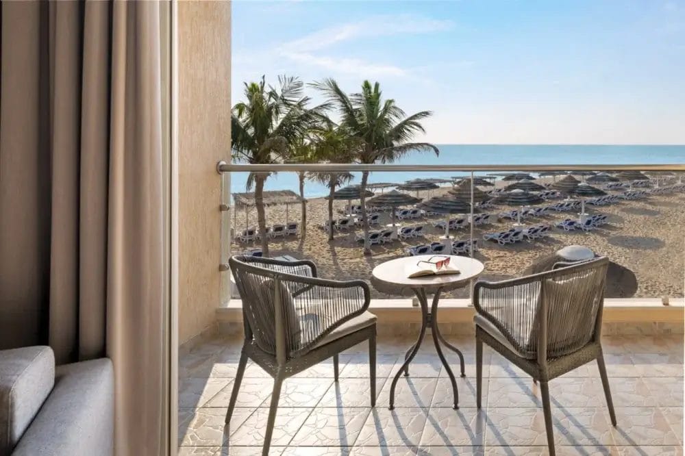 Un balcone con due sedie di vimini e un tavolino si affaccia su una spiaggia sabbiosa con palme, lettini, ombrelloni e un mare calmo e azzurro sotto un cielo limpido: l'ideale per una fuga rilassante a Bozza automatica.