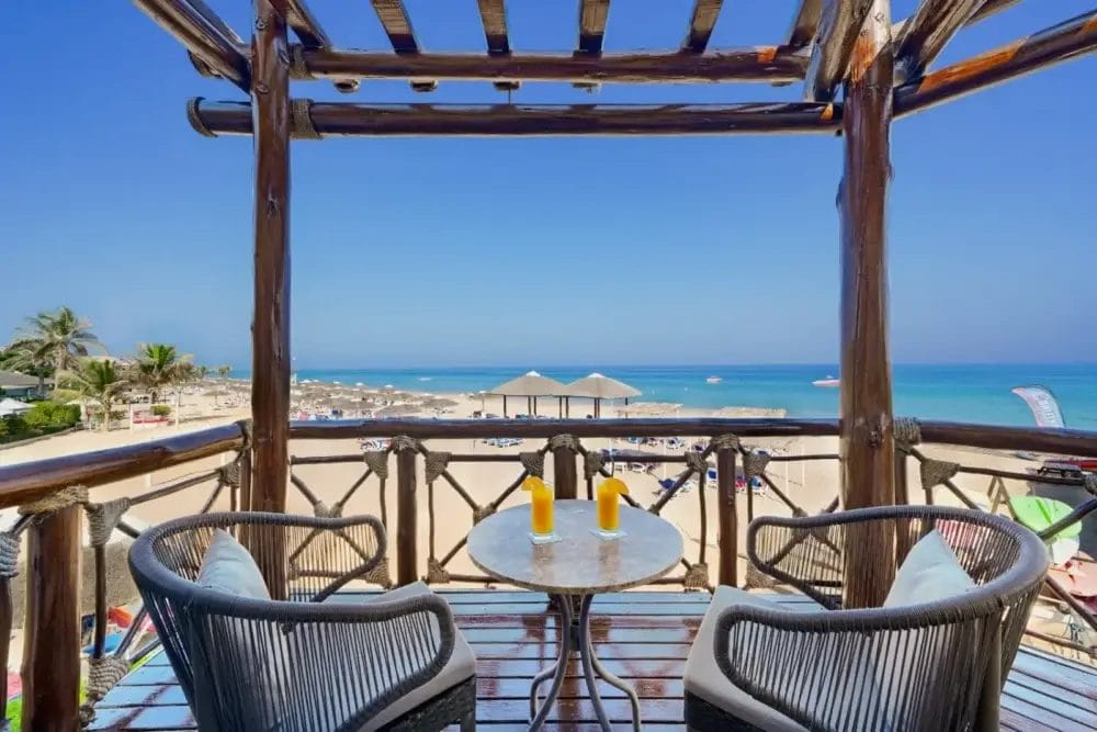 Un balcone sulla spiaggia con due sedie e un tavolino con due aranciate offre la vista perfetta di Bozza automatica: spiaggia sabbiosa, mare azzurro, lettini e ombrelloni sotto un cielo limpido.
