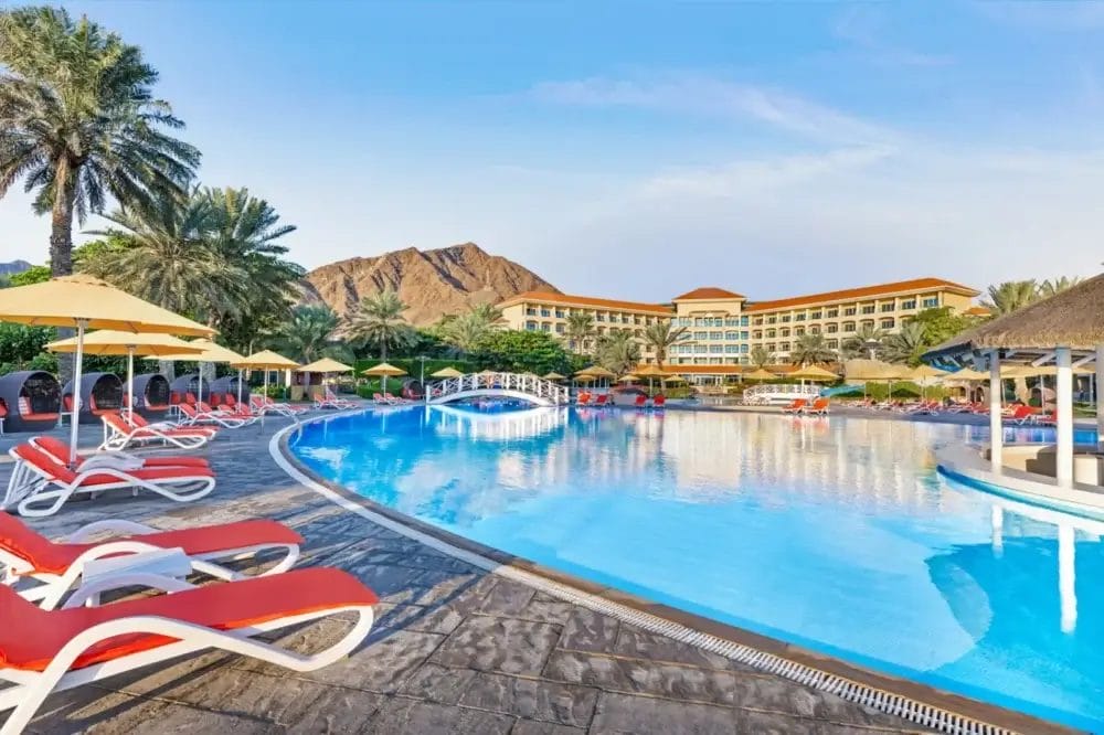 Una grande piscina all'aperto con sedie a sdraio rosse e bianche, ombrelloni gialli, palme e una bozza automatica. Un ponte attraversa la piscina, mentre un edificio del resort con balconi si erge sullo sfondo di una vista sulle montagne.