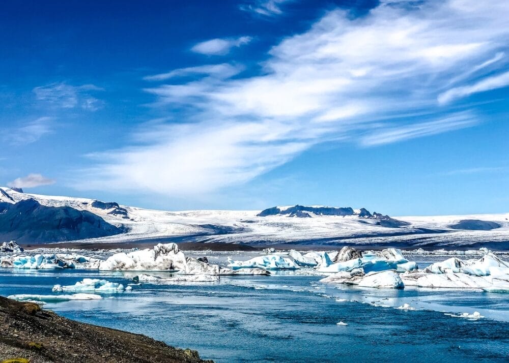 Grandi pezzi di ghiaccio galleggiano in una laguna glaciale blu sotto un cielo azzurro brillante con nuvole sparse, circondato da montagne innevate - una scena impressionante che sembra quasi una bozza automatica della natura.