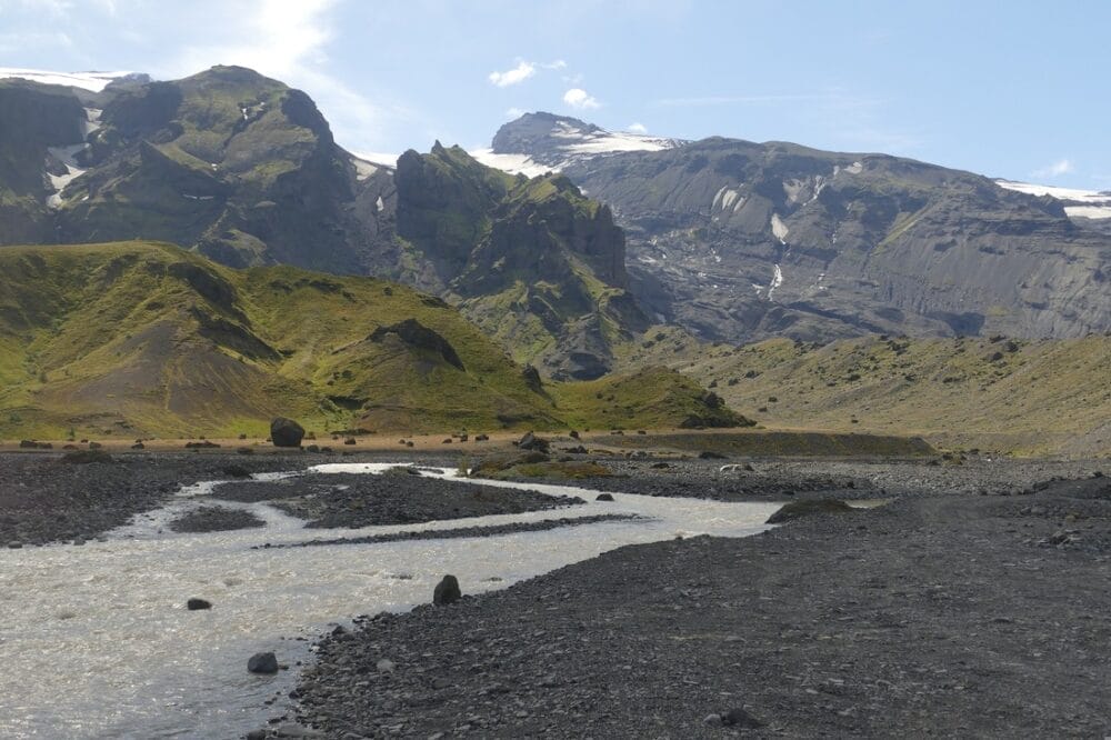 Un ampio fiume scorre in una valle rocciosa con colline verdi ricoperte di muschio, mentre montagne scure si ergono sullo sfondo sotto un cielo blu con nuvole sparse: una stupefacente bozza automatica dell'arte della natura.