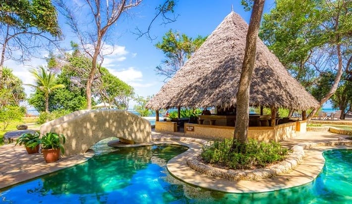 La scena di un resort tropicale sull'isola di Chale, in Kenya, è caratterizzata da una capanna con il tetto di paglia accanto a una piscina blu chiaro circondata da una vegetazione lussureggiante. Un piccolo ponte ad arco attraversa la piscina, con vista sull'oceano di The Sands sotto un cielo luminoso.