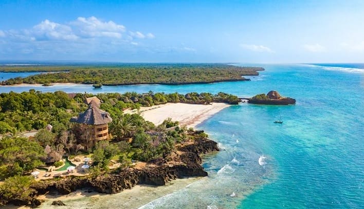 Veduta aerea dell'isola di Chale con una vegetazione lussureggiante, spiagge sabbiose, acque turchesi e un resort balneare con tetto di paglia vicino alla riva. Piccole onde si infrangono sulle rocce mentre la costa si incurva intorno al mare cristallino.