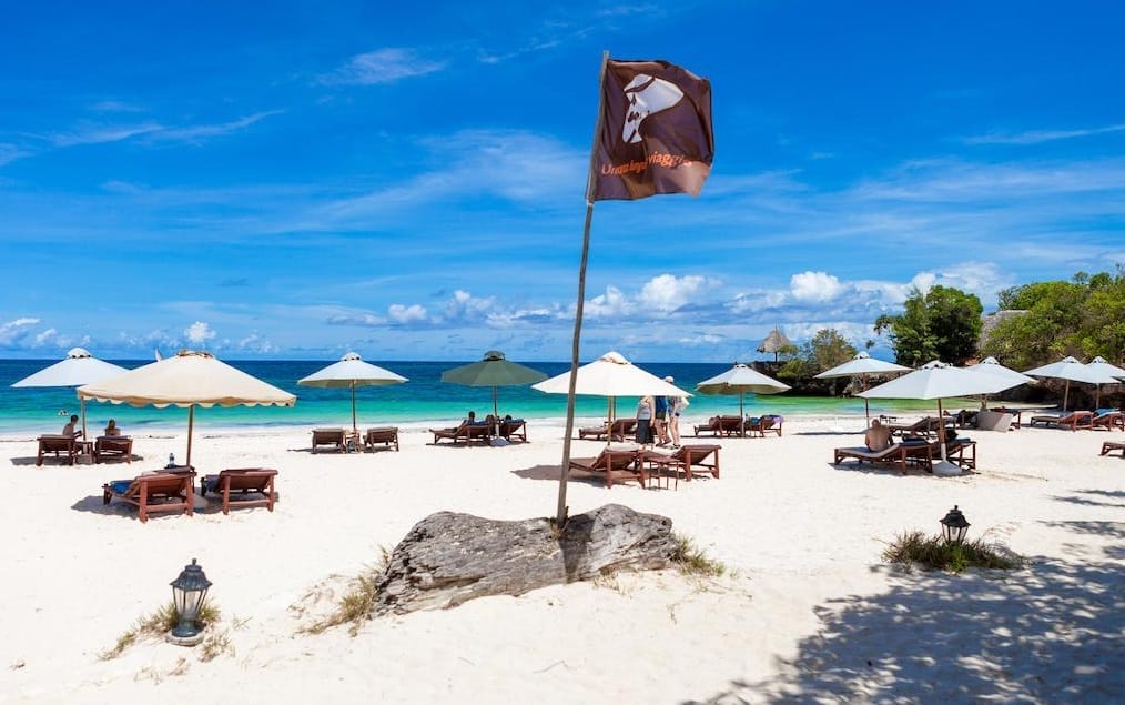 Spiaggia sabbiosa con sedie a sdraio e ombrelloni in legno a Sands At Chale Island, persone che si rilassano in riva all'oceano turchese, una bandiera marrone su un'asta al centro e un cielo blu brillante con nuvole sparse in alto in questo resort balneare del Kenya.