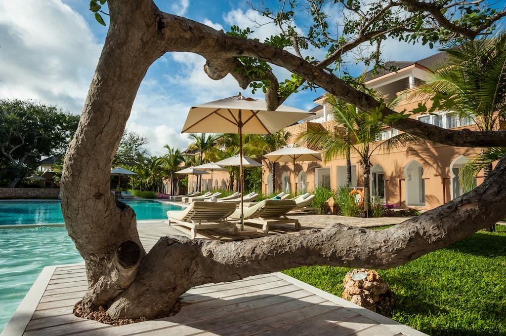 Un grande albero contorto incornicia una vista di sedie a sdraio a bordo piscina con ombrelloni bianchi accanto a una piscina blu chiaro nel resort di lusso Medina Palms a Watamu, con un edificio color pesca e palme verdi sotto un cielo parzialmente nuvoloso.