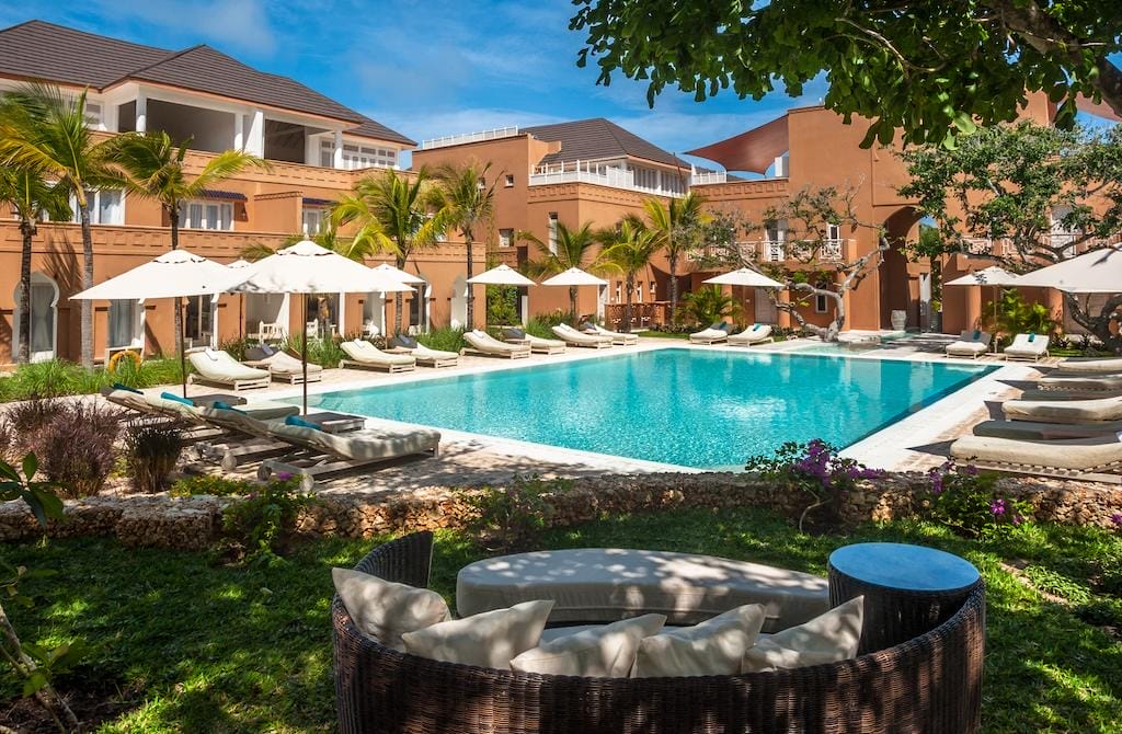 Il Medina Palms, un hotel di lusso in Kenya, dispone di un'ampia piscina all'aperto circondata da lettini e ombrelloni, con palme e un paesaggio lussureggiante, circondato da eleganti edifici color terracotta sotto un cielo blu.