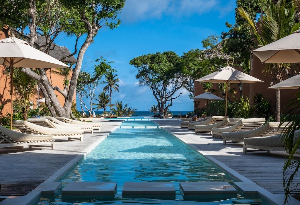 La lussuosa piscina all'aperto del Medina Palms, fiancheggiata da sedie a sdraio e ombrelloni, è circondata da alberi tropicali e dal verde, e conduce a una splendida vista sull'oceano sotto un cielo blu brillante.
