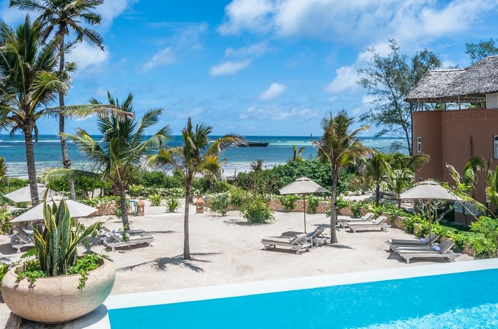 Un resort di lusso a Watamu, il Medina Palms vanta palme, lettini e ombrelloni sulla sabbia bianca, il tutto visto da una serena piscina. L'oceano e il cielo blu con nuvole sparse creano uno splendido scenario tropicale.