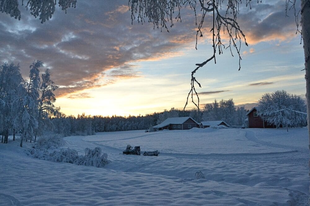 Paesaggio innevato all'alba, punteggiato da lontani fienili rossi e alberi smerigliati. Tracce disegnano la neve, mentre una Bozza automatica di nuvole si staglia nel cielo, illuminato da una luce calda che si spande all'orizzonte.