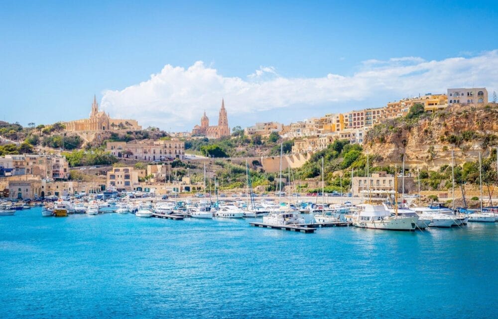 Un porto turistico pieno di barche e yacht sull'acqua blu brillante, circondato da edifici colorati e scogliere rocciose, presenta due grandi chiese con cupole e guglie sullo sfondo sotto un cielo parzialmente nuvoloso: una scena perfetta per una Bozza automatica.