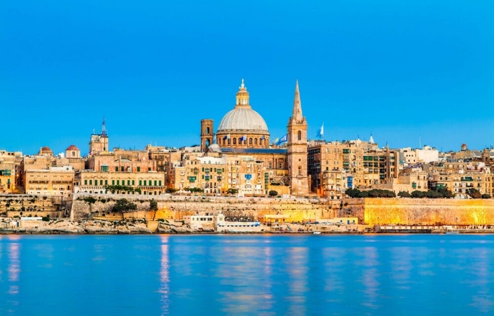 Una vista panoramica di La Valletta, a Malta, con edifici storici, una grande cupola e l'alta guglia di una chiesa, il tutto illuminato al tramonto e riflesso nelle calme acque azzurre del porto, immortalato in perfetto stile Bozza automatico.