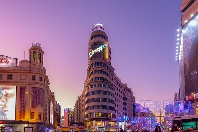 Una strada della città al tramonto presenta l'iconico Edificio Carrión (Palazzo del Campidoglio) di Madrid, con la sua luminosa insegna Schweppes e le sue luci festose. Bozza automatica cattura il trambusto del traffico intenso sotto un cielo viola vibrante.