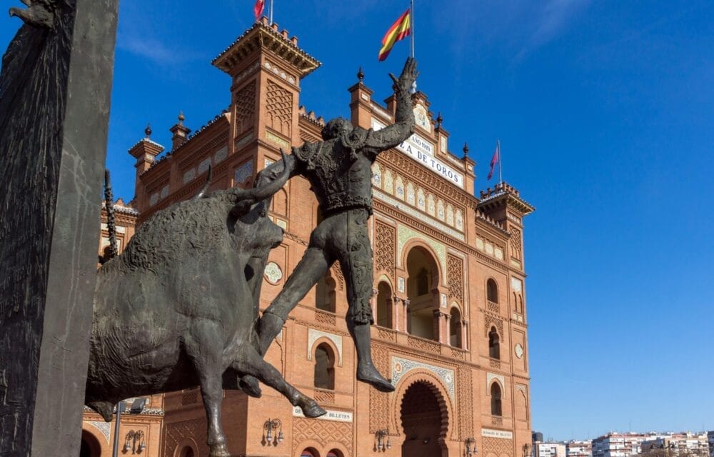 Una statua di bronzo raffigurante un torero e un toro si trova davanti all'arena Las Ventas di Madrid, in mattoni rossi. Le bandiere spagnole sventolano in cima all'edificio ornato sotto un cielo azzurro e limpido, a simboleggiare la vibrante cultura di Madrid.