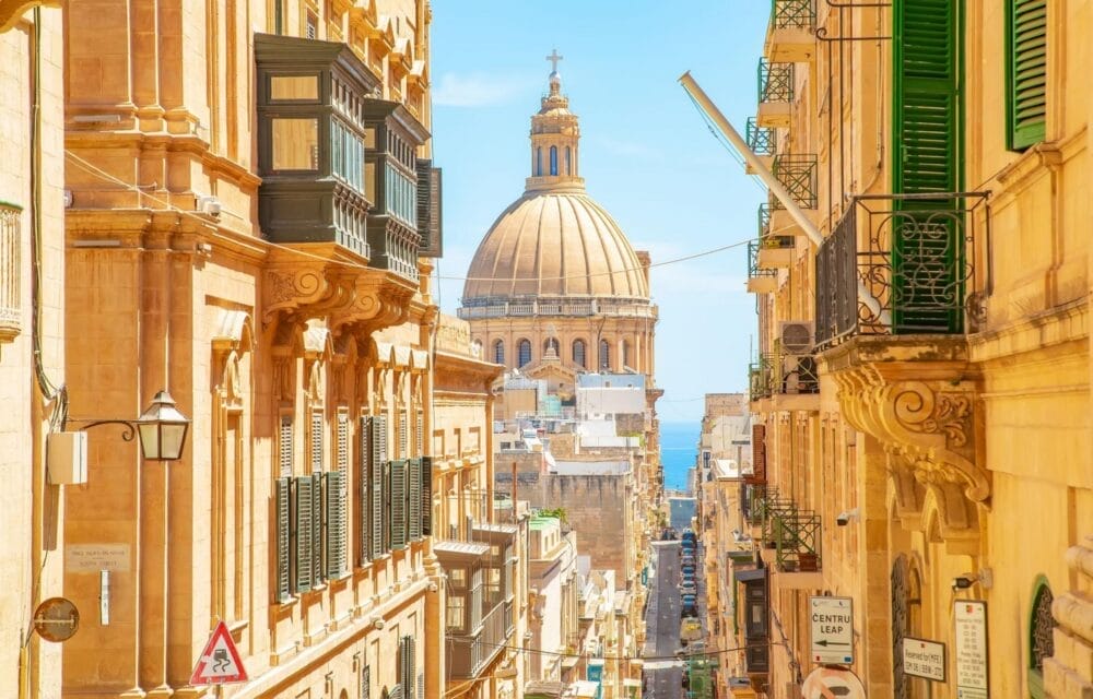 Una strada soleggiata di La Valletta, a Malta, fiancheggiata da edifici tradizionali in pietra gialla e balconi verdi, conduce alla cupola prominente di una chiesa. Bozza automatica si inserisce delicatamente nella vivace scena, con il mare blu visibile sullo sfondo.