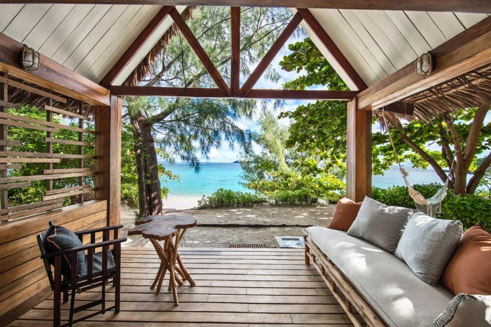 Patio in legno all'aperto con divano, tavolo rustico e sedia, affacciato su una spiaggia sabbiosa e sull'oceano cristallino del resort di lusso dell'isola malgascia Constance Tsarabanjina, circondato da alberi verdeggianti sotto un tetto parzialmente coperto.
