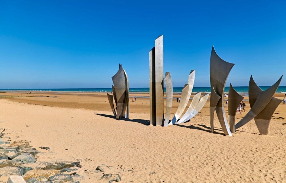 Una scultura dalle forme metalliche alte e astratte si erge su una spiaggia sabbiosa in Normandia sotto un cielo azzurro e limpido, con l'oceano sullo sfondo e le persone che passeggiano nelle vicinanze durante il loro tour di Capodanno.