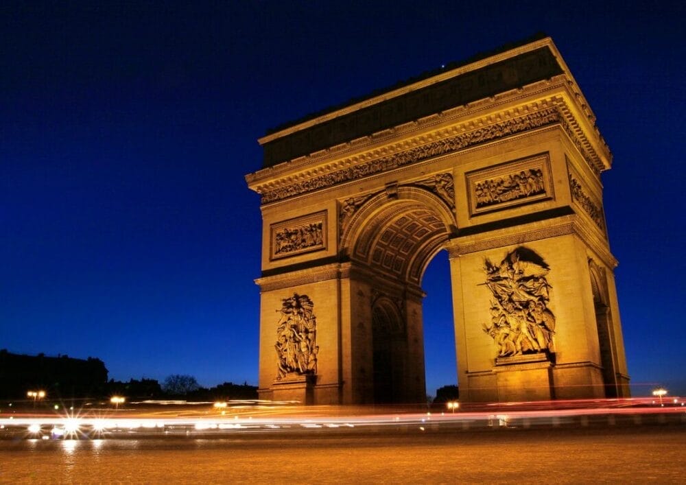 L'Arco di Trionfo di Parigi è illuminato di notte contro un cielo blu intenso, con scie luminose sfocate dei veicoli in transito alla sua base: un'accattivante bozza automatica di energia cittadina e architettura senza tempo.