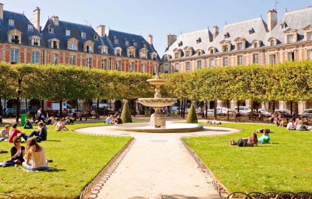 Le persone si rilassano sull'erba e sulle panchine intorno a una fontana centrale in pietra in un parco soleggiato di Parigi, con file di alberi ben curati ed edifici storici francesi sullo sfondo.