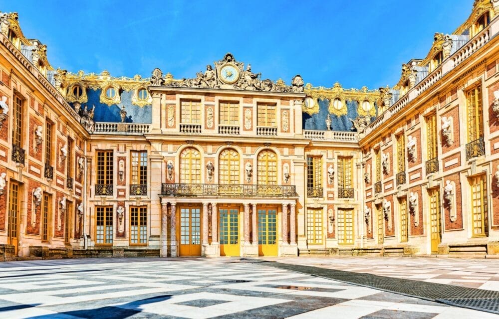 L'immagine mostra l'esterno ornato della Reggia di Versailles, con decorazioni dorate, grandi finestre e un'architettura simmetrica sotto un cielo blu brillante: una tappa iconica del tour di Capodanno in Normandia. Il cortile è caratterizzato da un pavimento in marmo a motivi.